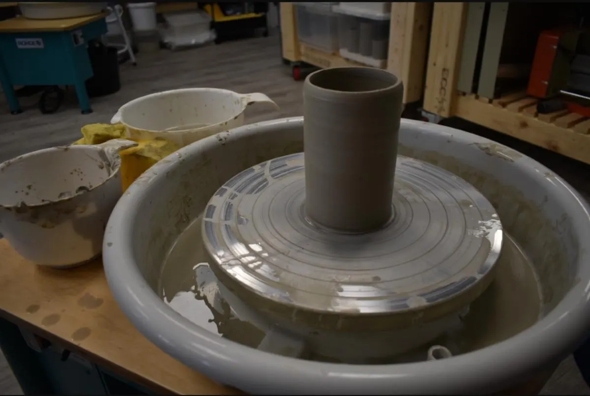 A cylindrical clay vessel being formed on a pottery wheel in a studio workshop.