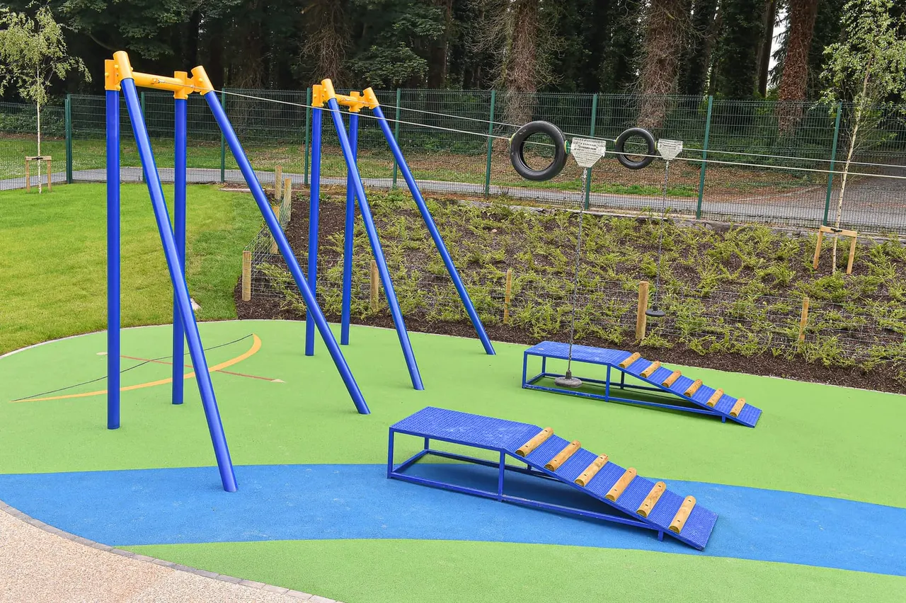 Outdoor play area with blue and yellow climbing frames, ramps and tyre obstacle course on coloured rubber matting.