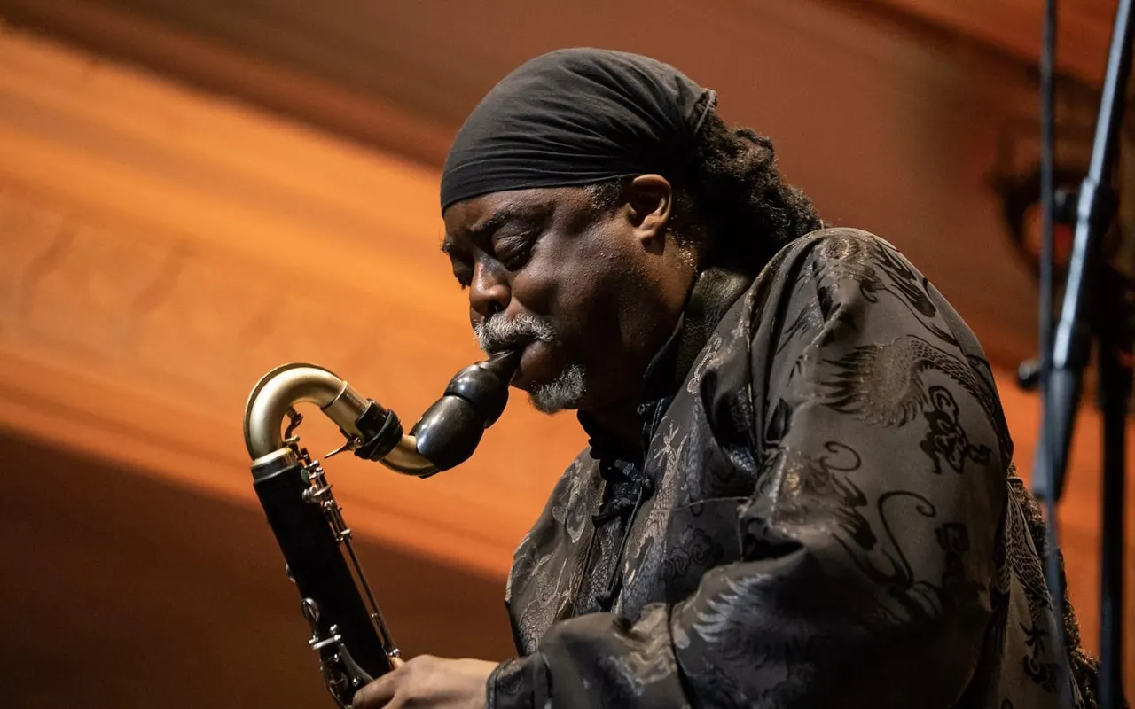 Man with beard and headwrap playing saxophone on stage against orange backdrop.