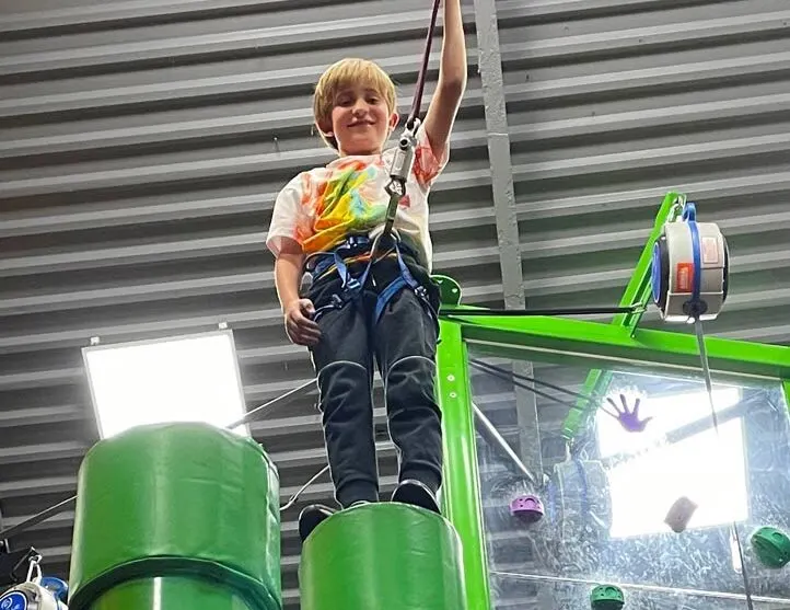 Young child suspended in the air whilst harnessed to a rope climbing activity, smiling with arm raised.