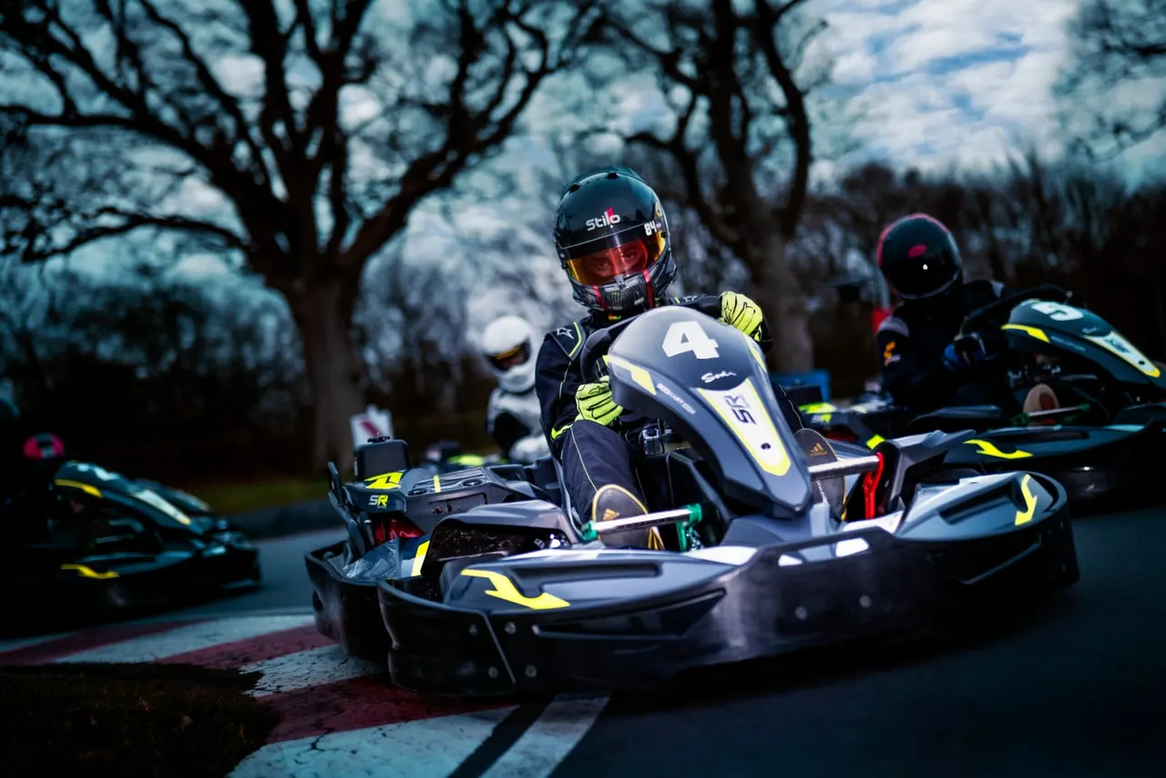 Kart racer in helmet and yellow gloves driving numbered go-kart on track with bare trees and hills beyond.