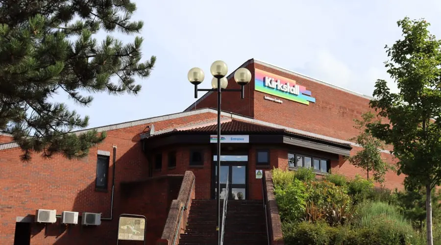 Exterior of a brick leisure centre building with an entrance sign and lamp post, surrounded by trees.