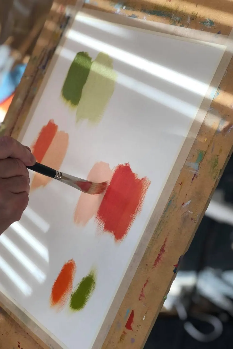 Person's hand holding paintbrush, painting coloured brushstrokes on white paper with red and green paint.