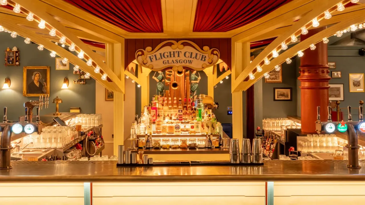 Ornate art deco bar interior with illuminated back bar displaying spirits bottles, brass taps and theatrical red and gold décor.