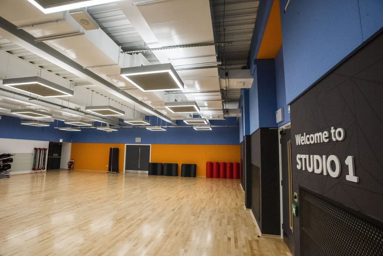 Empty fitness studio with wooden floor, blue and orange walls, and stacked equipment along the back wall.