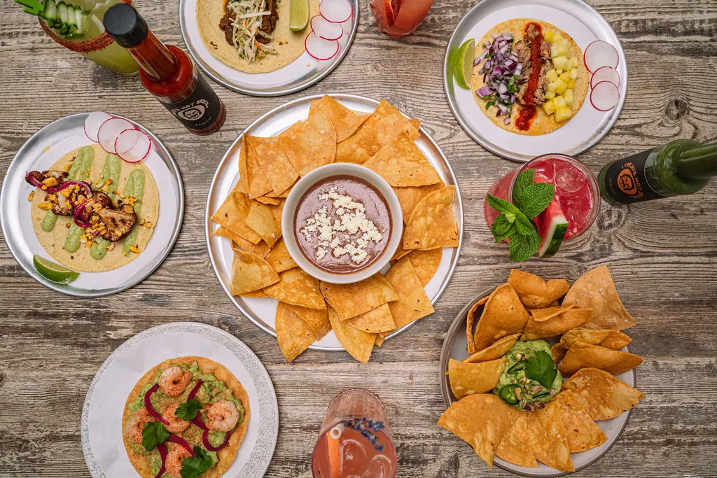 Overhead view of Mexican dishes including tacos, tortillas with toppings, tortilla chips with dip, and cocktails on a wooden table.