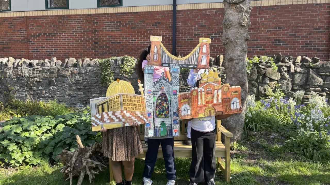 Three children wearing colourful illustrated landmark costumes stand in a garden with stone walls and flowering plants.
