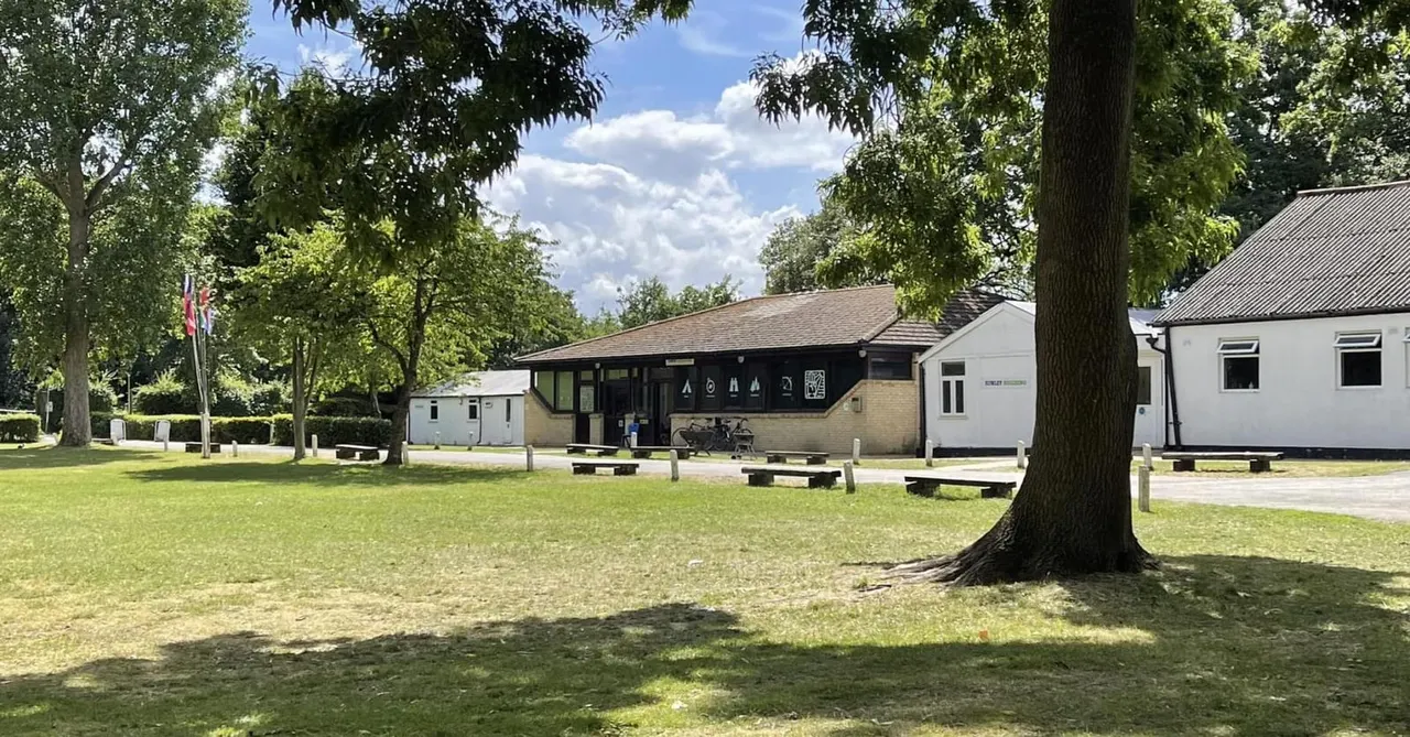 A leafy park area with mature trees, green lawn, picnic benches, and modern single-storey buildings under blue sky with white clouds.