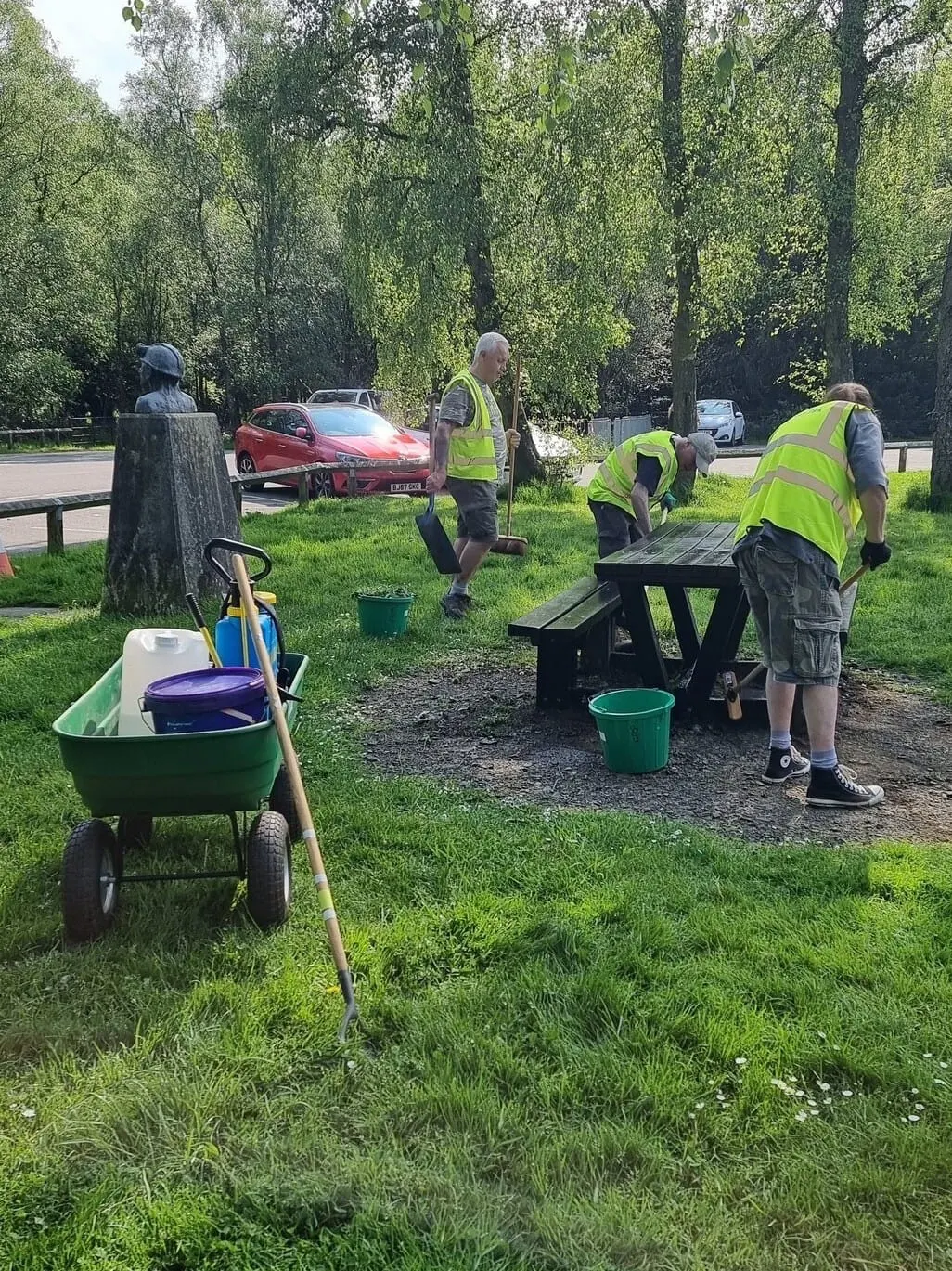 Workers in high-visibility jackets maintain a picnic area beside trees and parked cars.