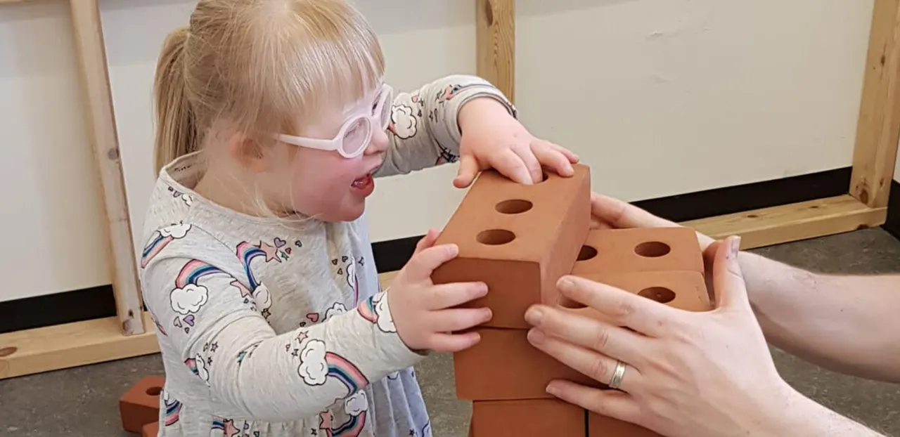 Young child wearing pink goggles explores terracotta building blocks with an adult during a sensory play activity.