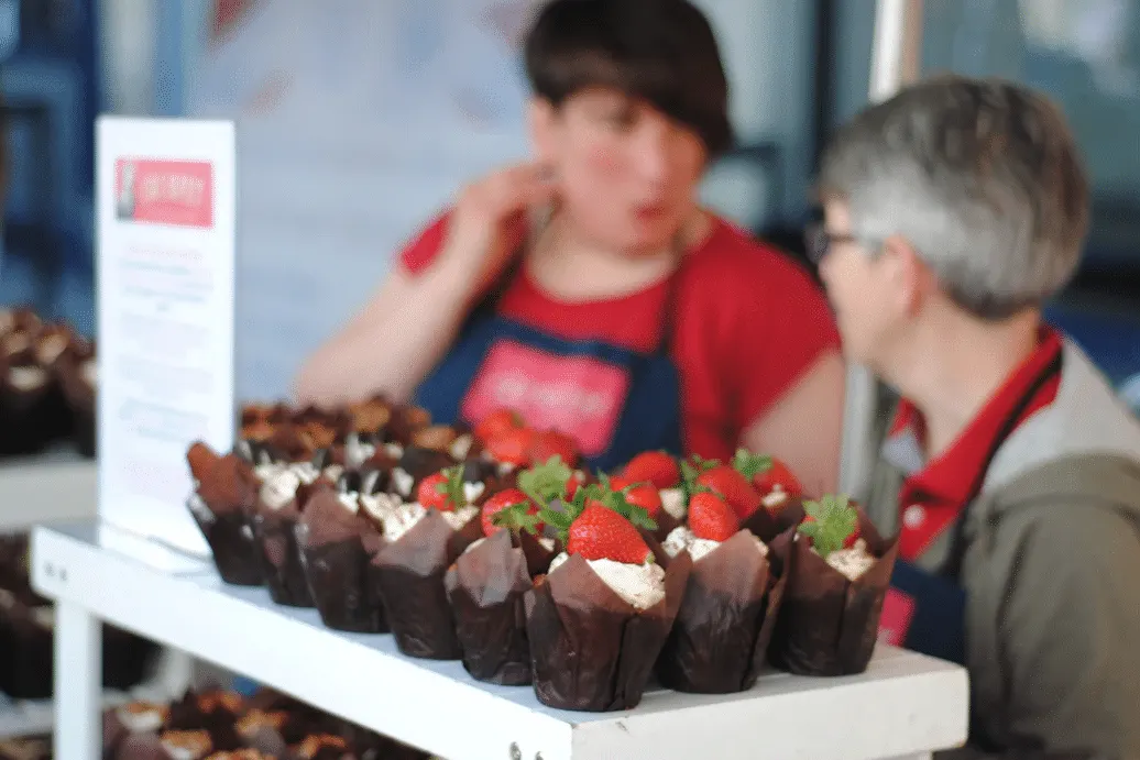 Chocolate desserts topped with strawberries displayed on a table at a food stall, with staff in red aprons behind.