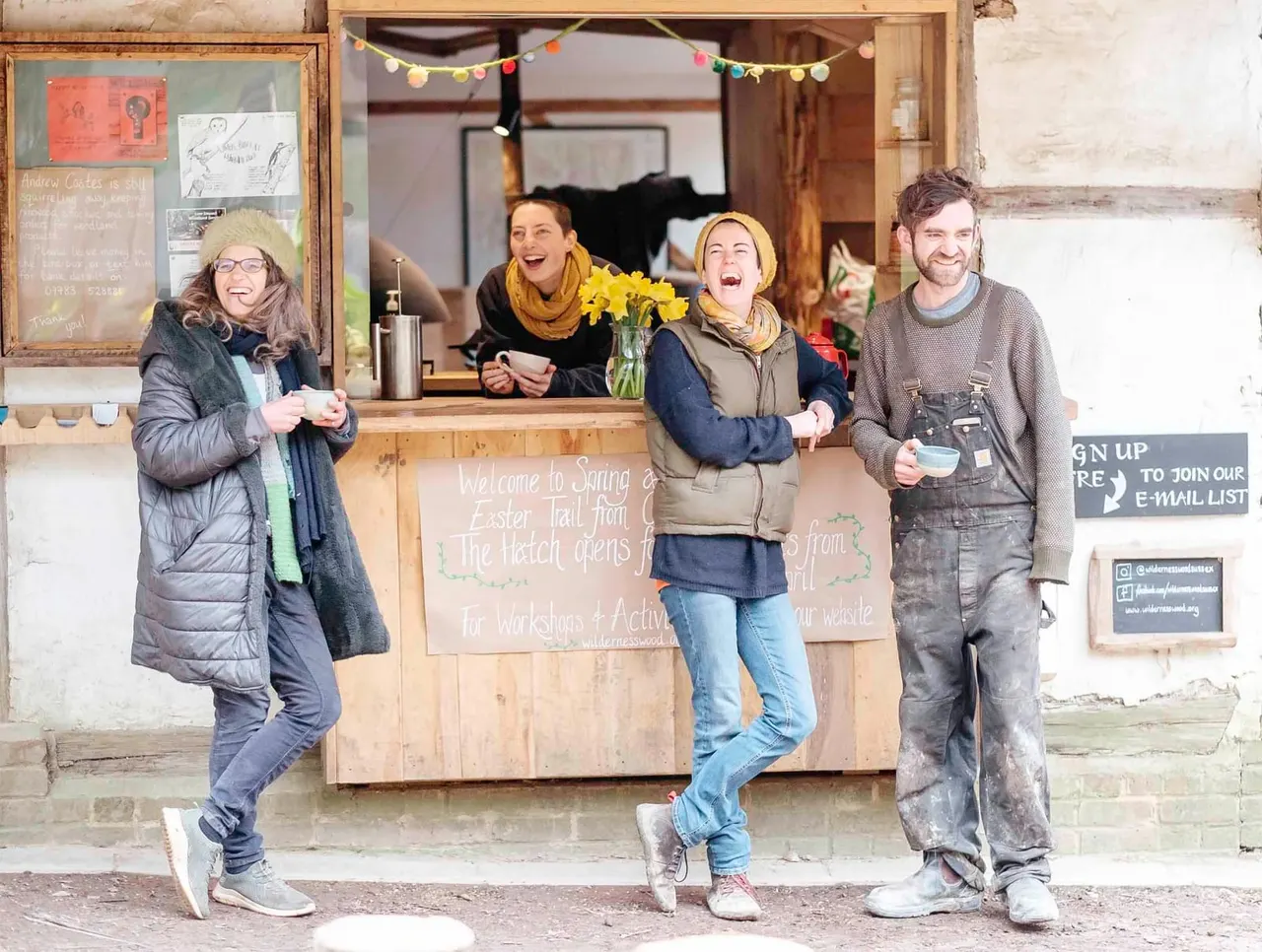 Four smiling people holding hot drinks outside a wooden refreshment hut decorated with colourful bunting.
