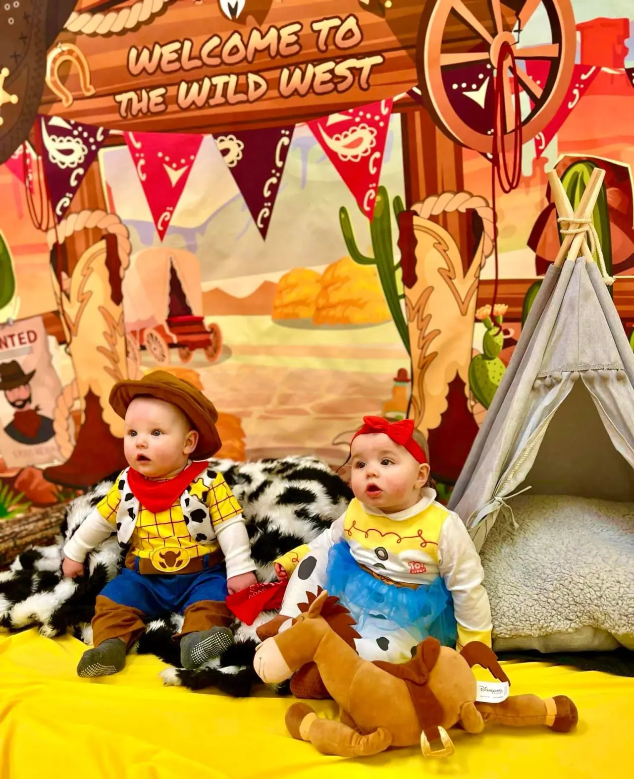 Two toddlers dressed as Wild West characters sit in front of a themed backdrop with a teepee and decorative props.