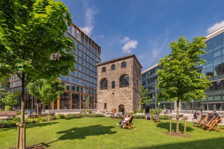 People relaxing in deckchairs on a grass courtyard surrounded by historic stone and modern buildings under blue sky.