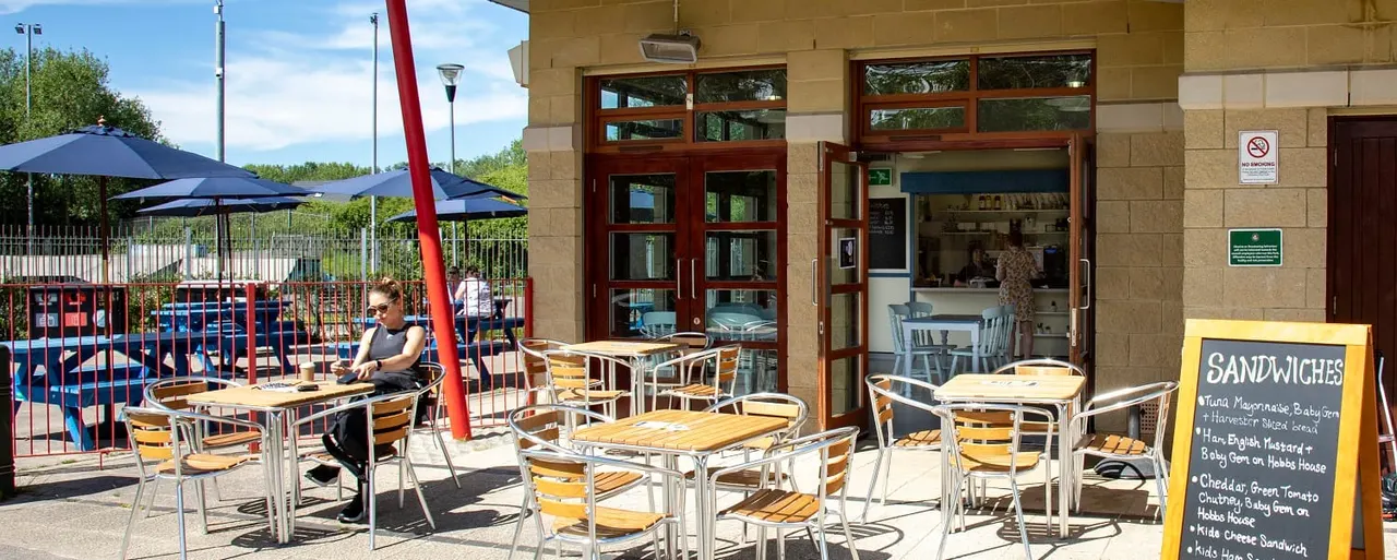 Outdoor café seating area at a leisure venue with tables and chairs, a person seated dining, building entrance with menu board, and children's play area visible beyond fencing.