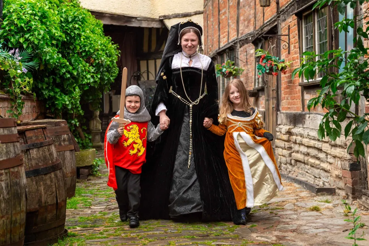 Woman in Tudor costume walking hand-in-hand with two children in period dress through a cobbled courtyard flanked by historic buildings.