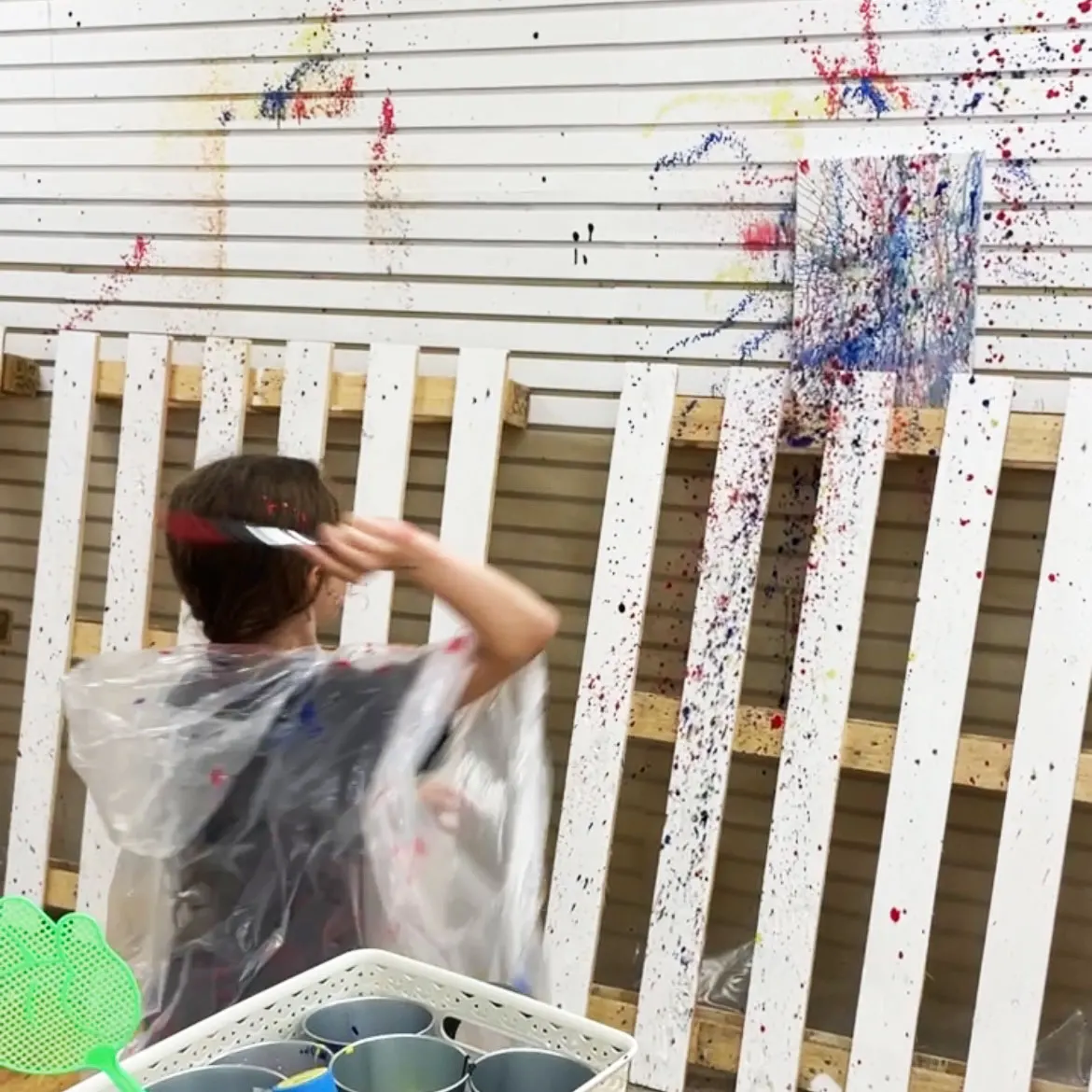 Child in protective apron splatter-painting a wooden frame covered in colourful paint splatters.