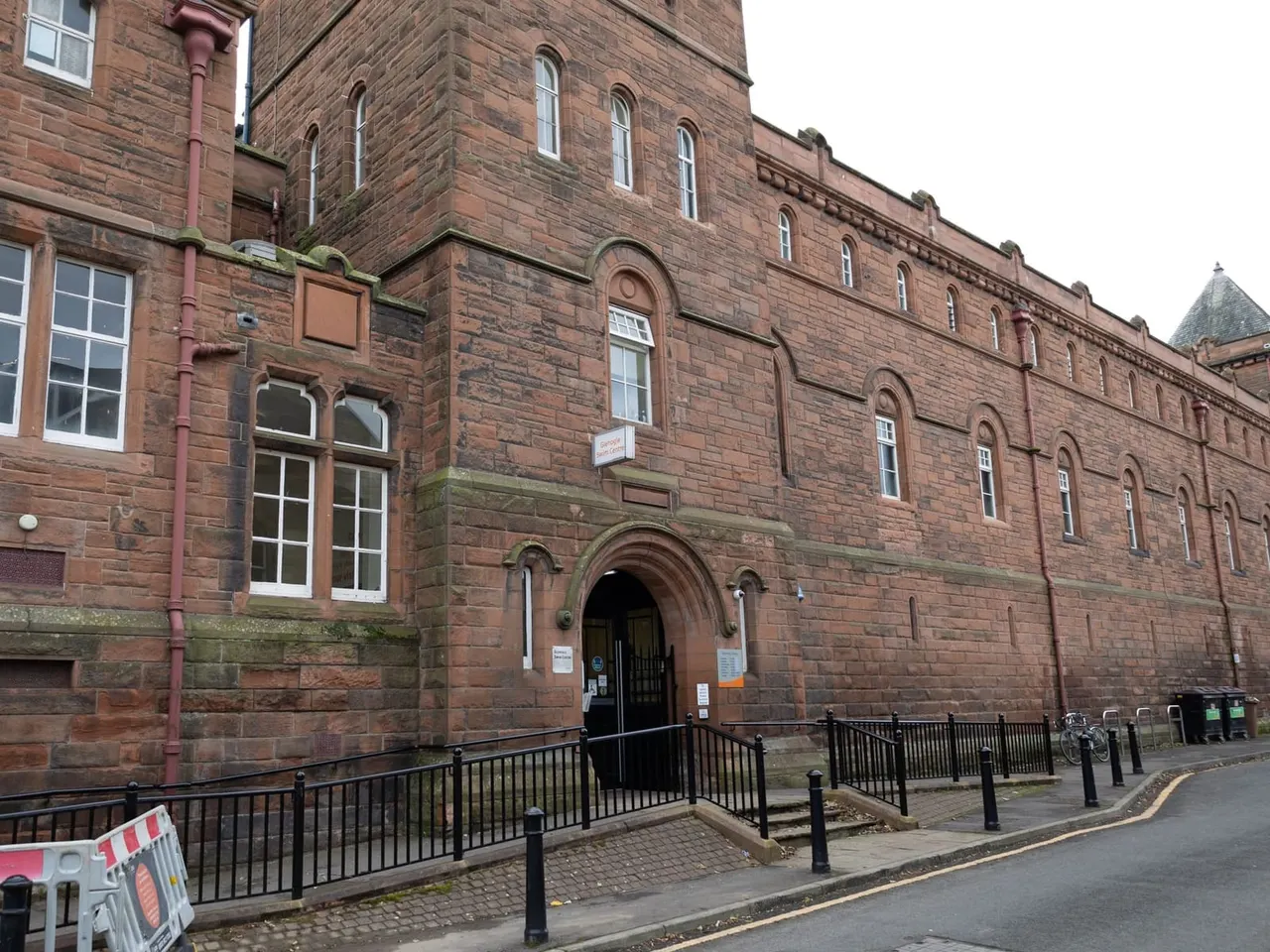Historic red sandstone building with arched entrance, tall windows and iron railings at street level.