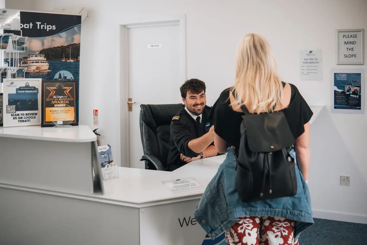 Staff member in uniform greets customer at reception desk of boat trips venue.