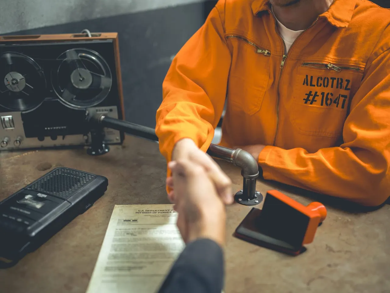 Person in orange prison-style jumpsuit operating vintage recording equipment at a desk with documents and devices.