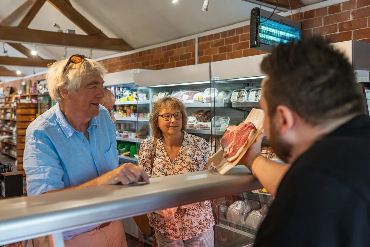 Customers at a deli counter speaking with staff members in a farm shop with refrigerated display cases.