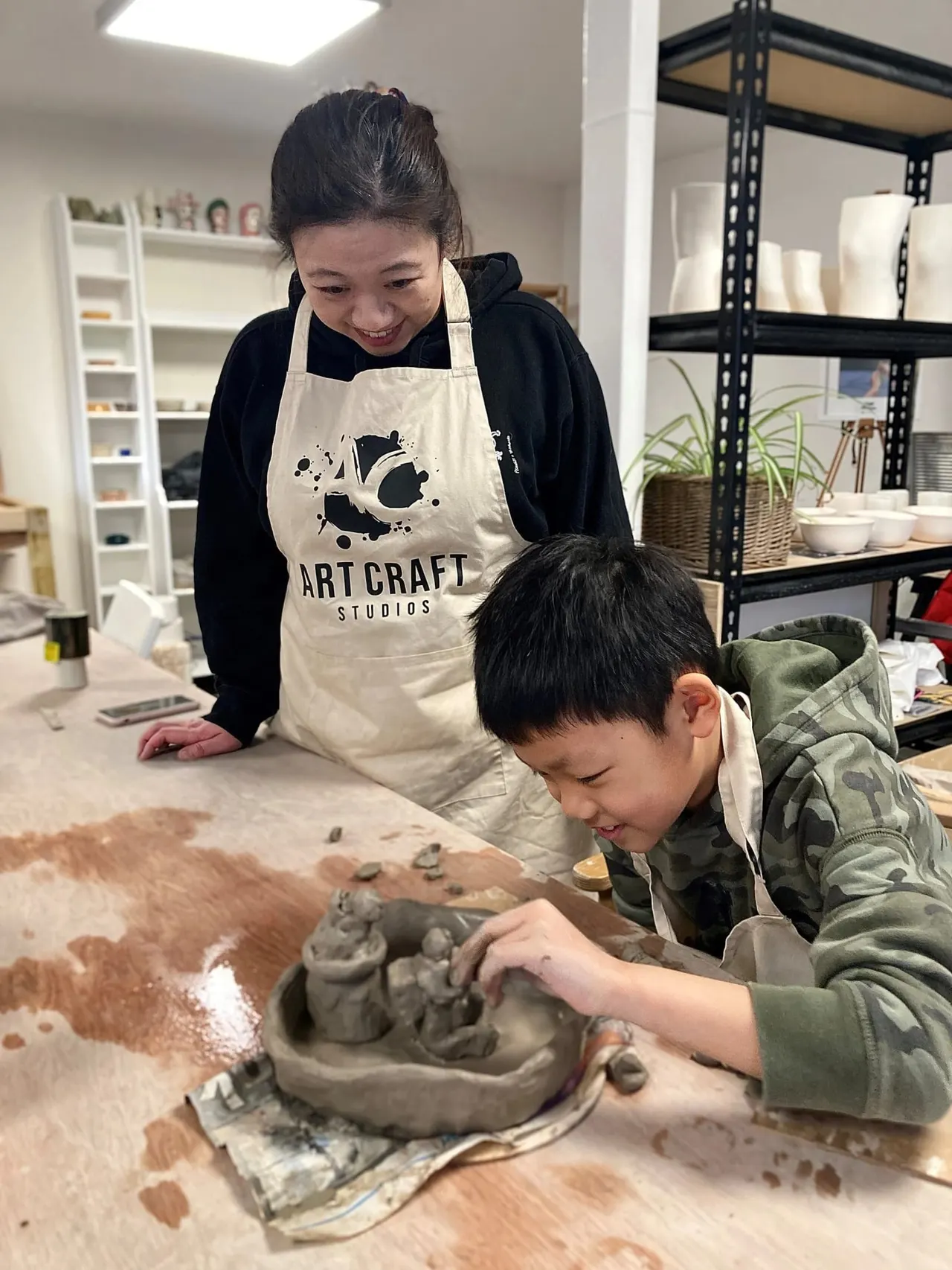 An adult and child work together on a clay sculpture at a pottery studio workbench.