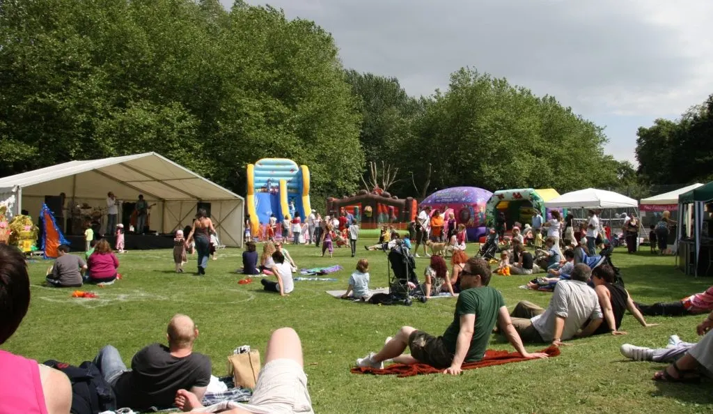 Outdoor family event with visitors relaxing on grass surrounded by inflatable attractions, marquees and trees on a sunny day.
