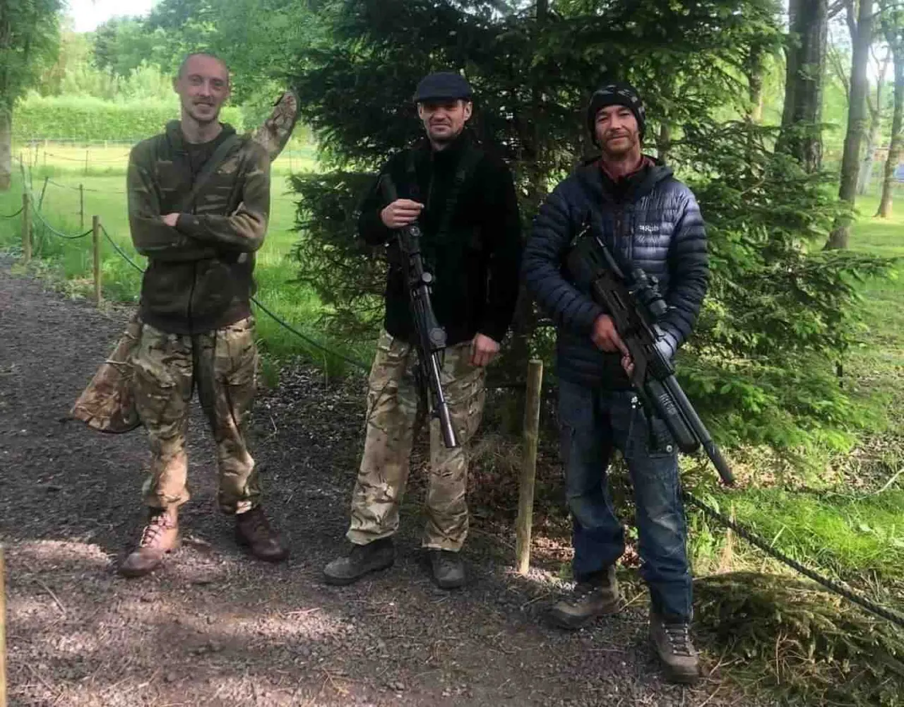 Three men holding rifles stand on a woodland path with green fields and trees behind them.