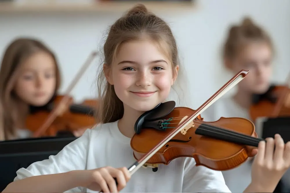Girl in white shirt smiling while playing violin, with two other young musicians holding violins in the background.