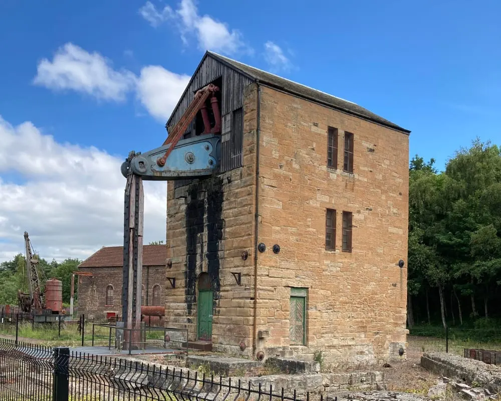 Historic red sandstone colliery winding engine house with vintage pit machinery displayed outside, set against blue sky.