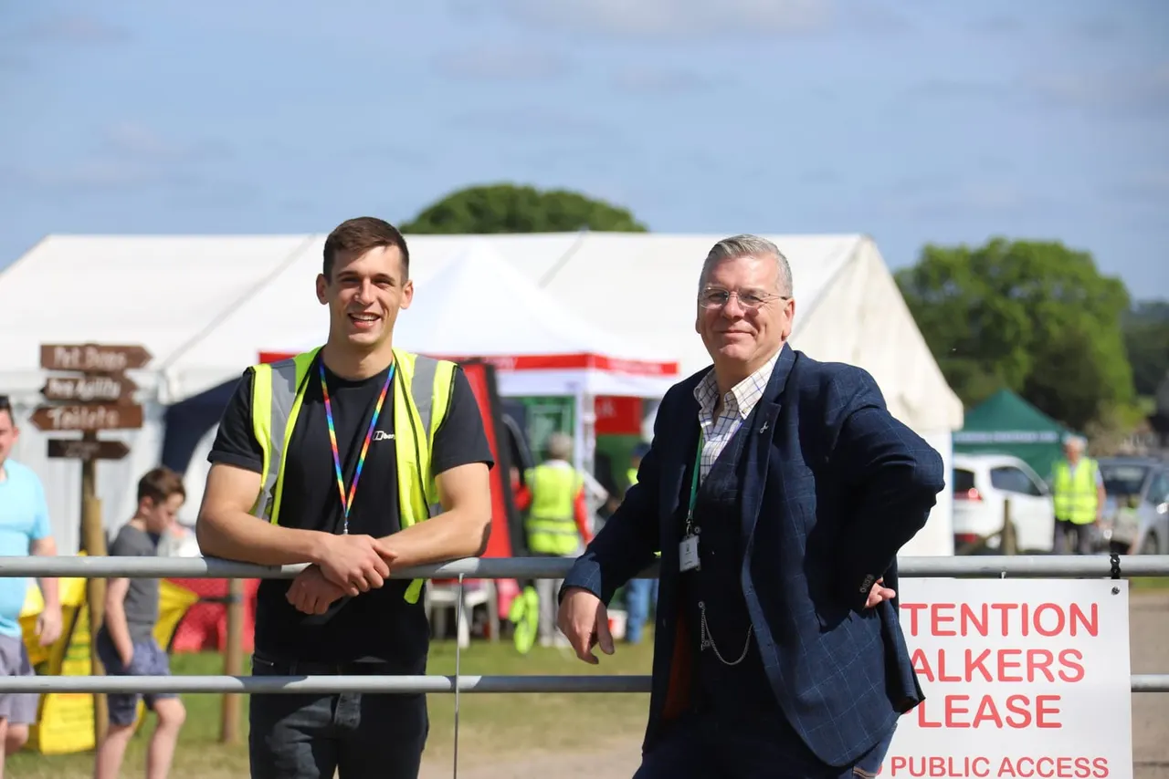 Two men stand at a railing at an outdoor event, with a large white marquee and trees visible behind them.