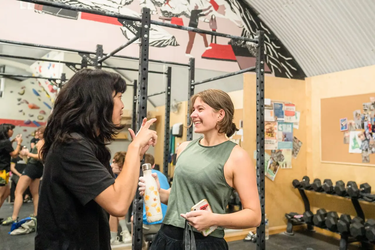 Two women chat in a gym with climbing wall, exercise equipment and dumbbells visible.