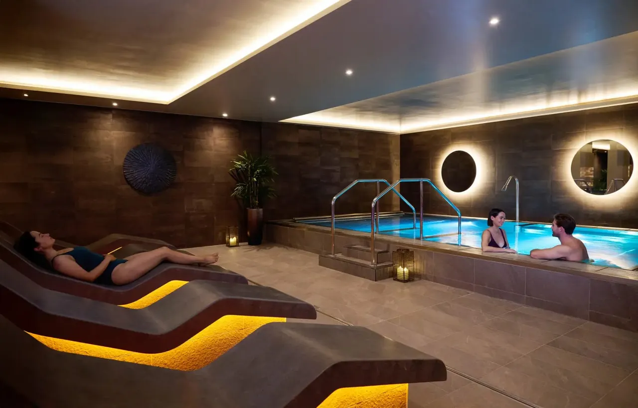 Guests relax on illuminated loungers and swim in a brightly lit indoor pool with contemporary design features.