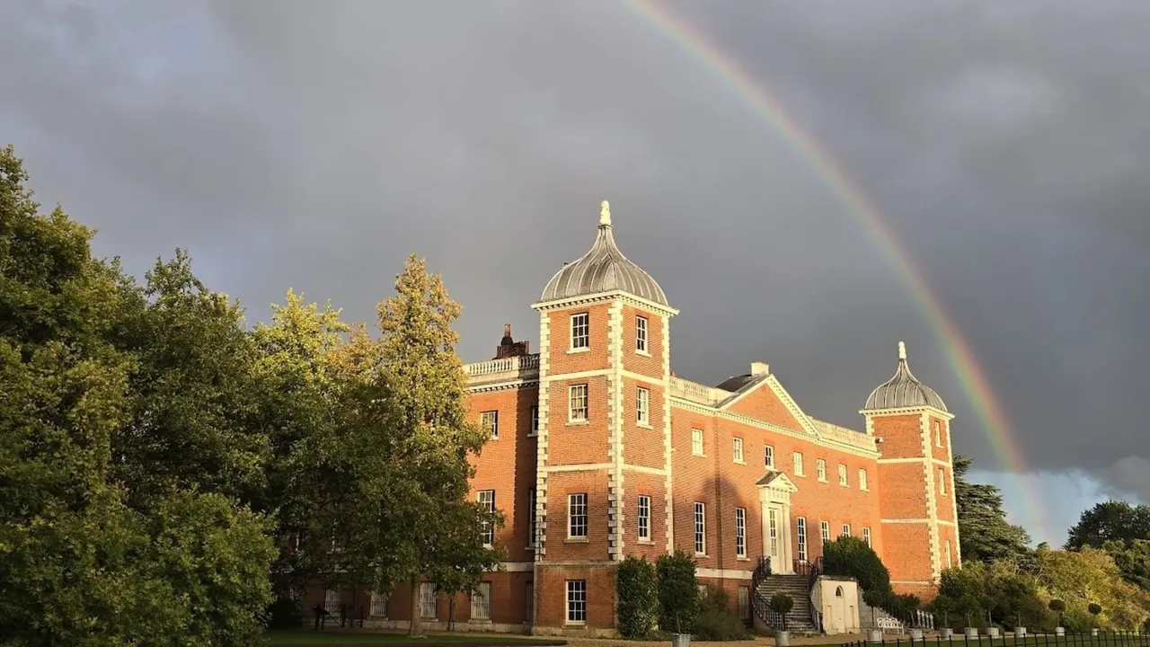 Red brick Georgian country house with domed turrets beneath a dramatic rainbow and stormy sky.
