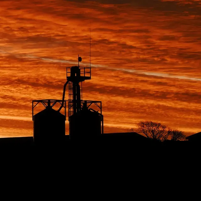 Silhouette of grain storage silos and a communications tower against a dramatic orange sunset sky.