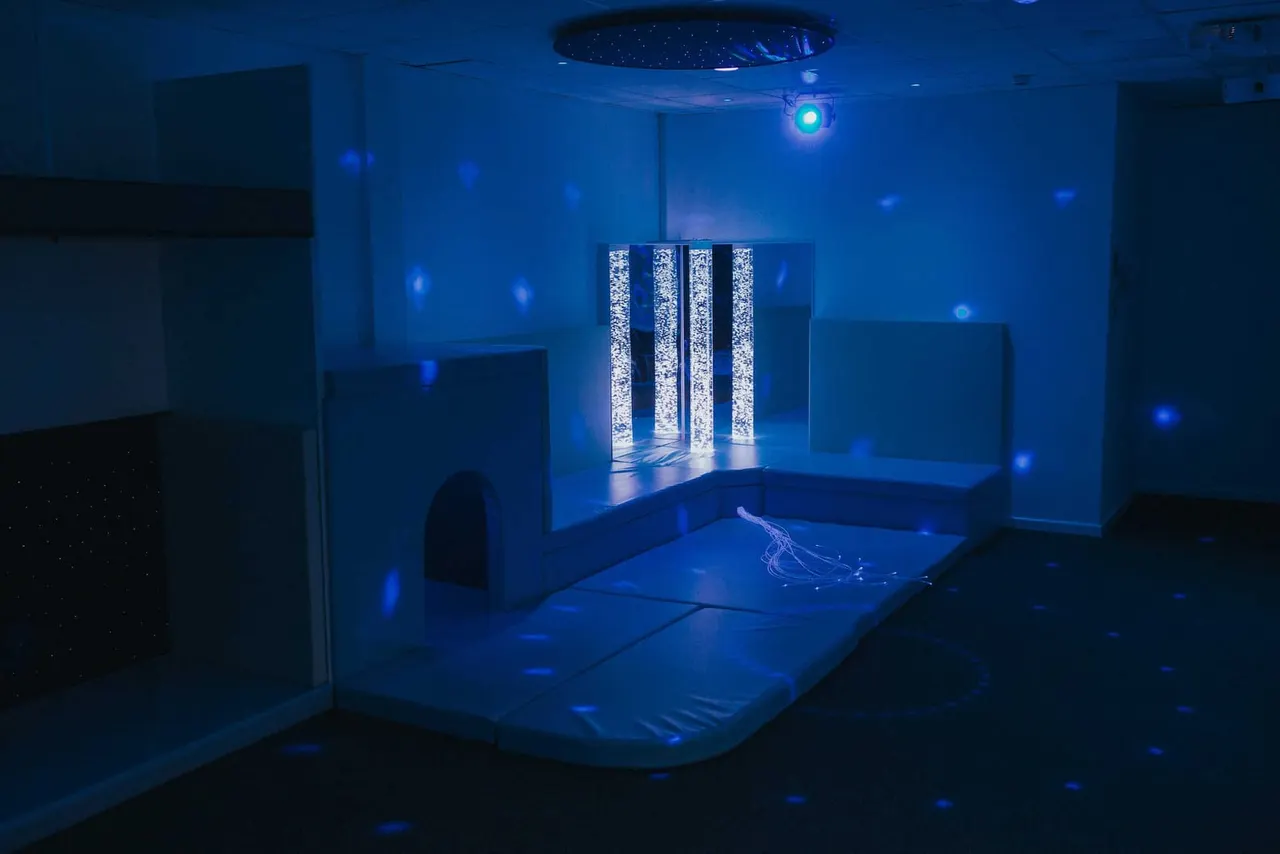 A darkened interactive play space with blue lighting, illuminated bubble columns, and sensory installations for children.