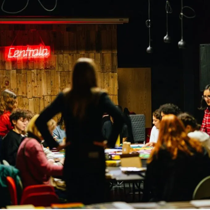 A person leads a creative activity or workshop at a table surrounded by seated participants in a modern venue with neon signage.
