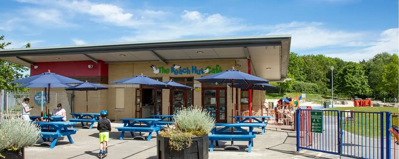 Child on scooter at outdoor café with blue picnic tables, umbrellas and playground equipment visible.