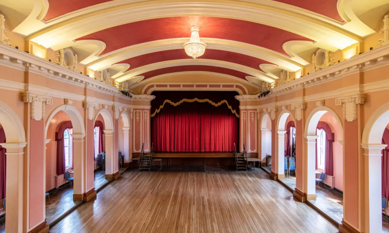 Empty theatre or assembly hall interior with ornate ceiling, red stage curtains and arched architectural details.