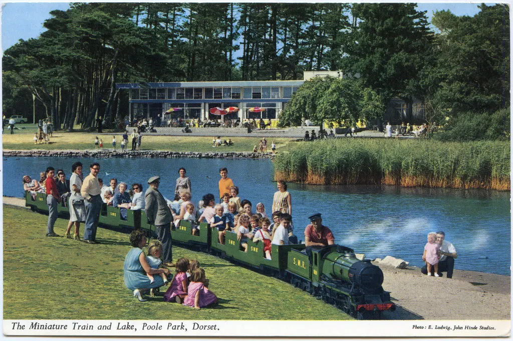 A miniature train filled with passengers travels along a lakeside track while visitors enjoy the grassy waterfront and modern pavilion building nearby.