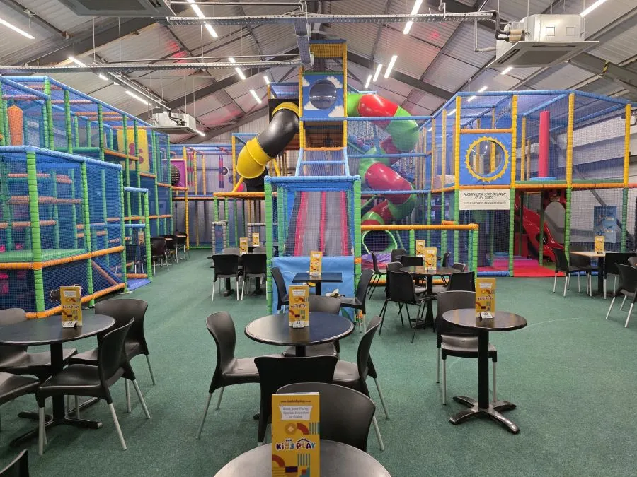 Indoor play centre with colourful climbing structures, slides and tunnels, seating tables in foreground, and children playing.