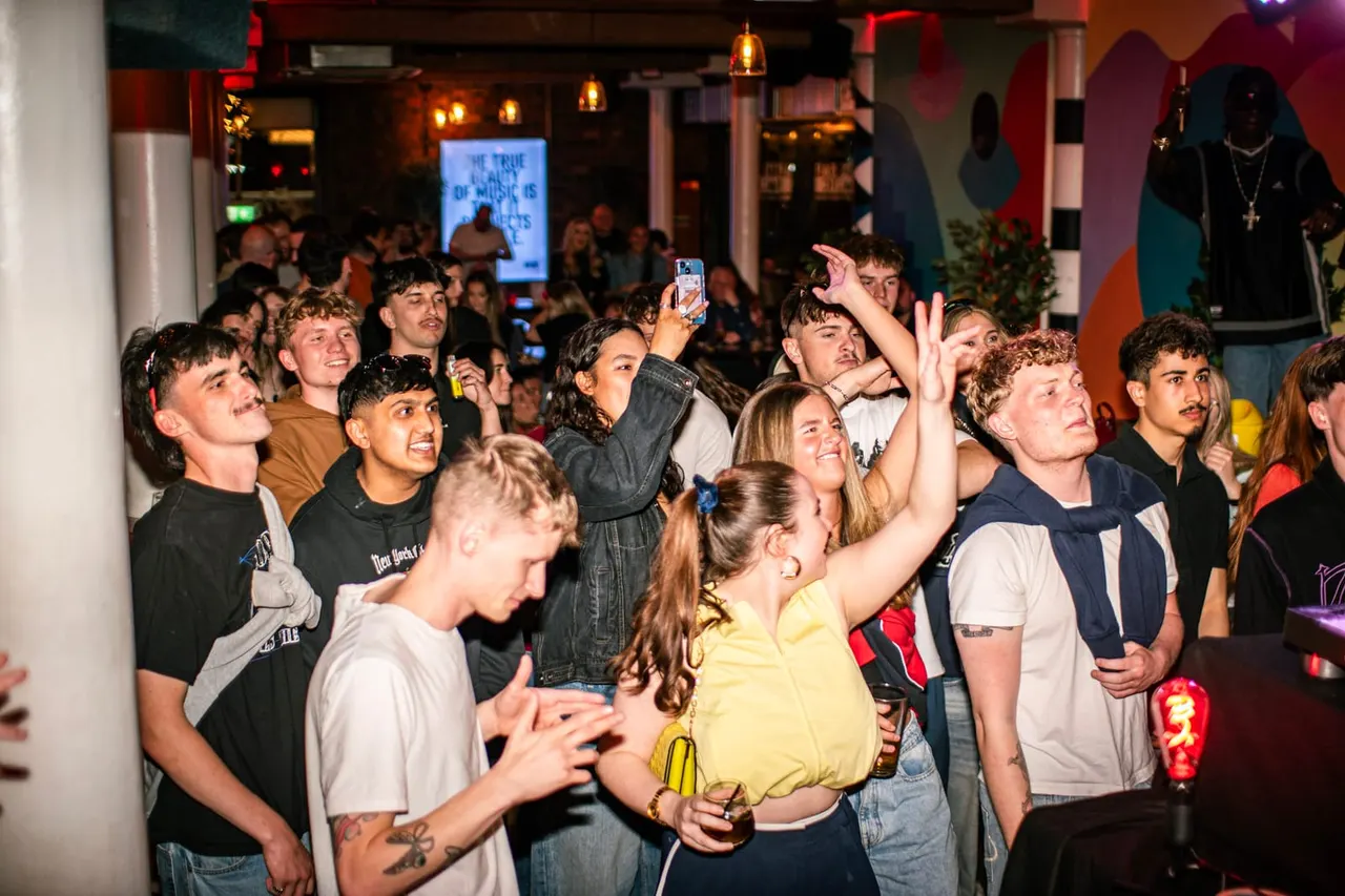 Crowd of young people dancing and watching a performance at an indoor nightclub venue with colourful lighting.