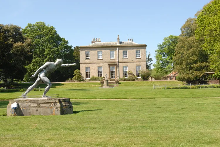 Stone sculpture of a figure in diving pose on manicured lawn with Georgian stately home and wooded grounds beyond.