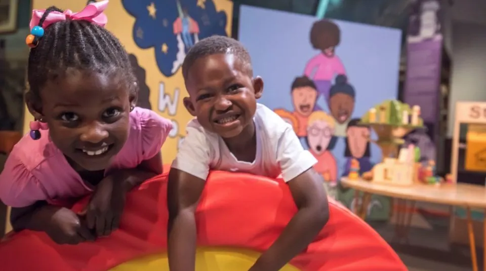 Two young children smiling at the camera whilst playing on a large red and yellow cushion in a colourful indoor play area.