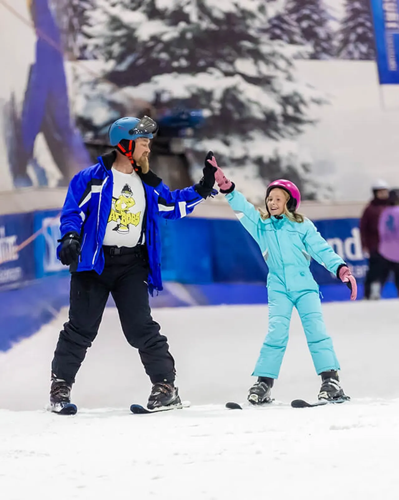 Adult instructor and young child ice skating together indoors, holding hands on an ice rink.