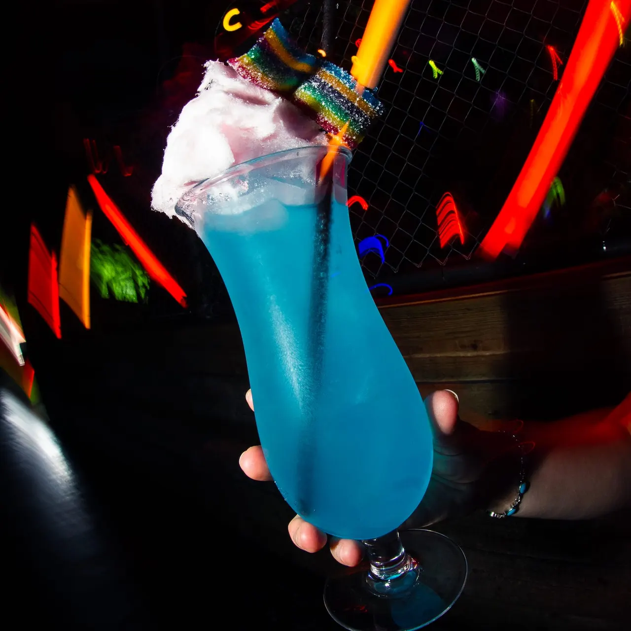 Hand holding a bright blue cocktail topped with white foam and rainbow glitter rim, surrounded by colourful neon lights.
