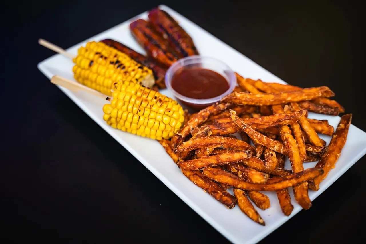 Grilled corn cobs, charred plantain slices, and sweet potato fries served with dipping sauce on a white plate.