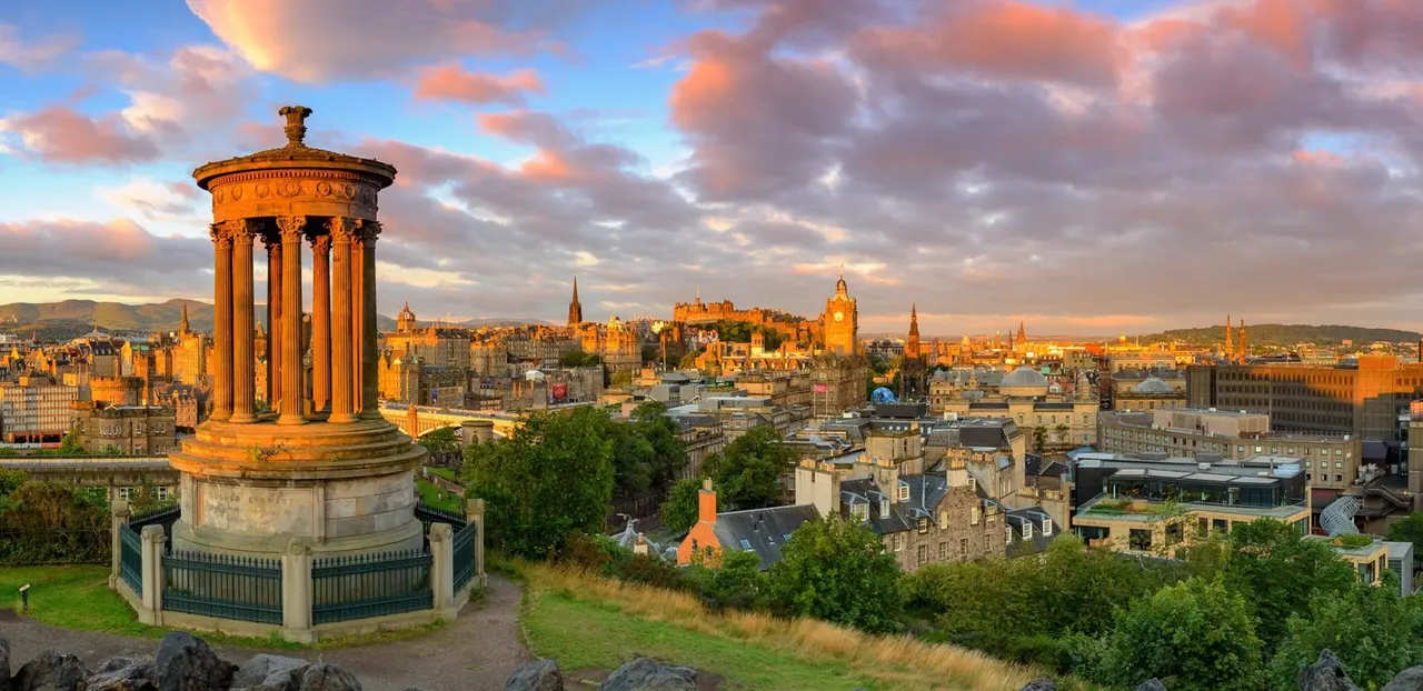 A classical columned monument overlooks Edinburgh's cityscape at golden hour, with historic buildings and spires visible across the skyline.
