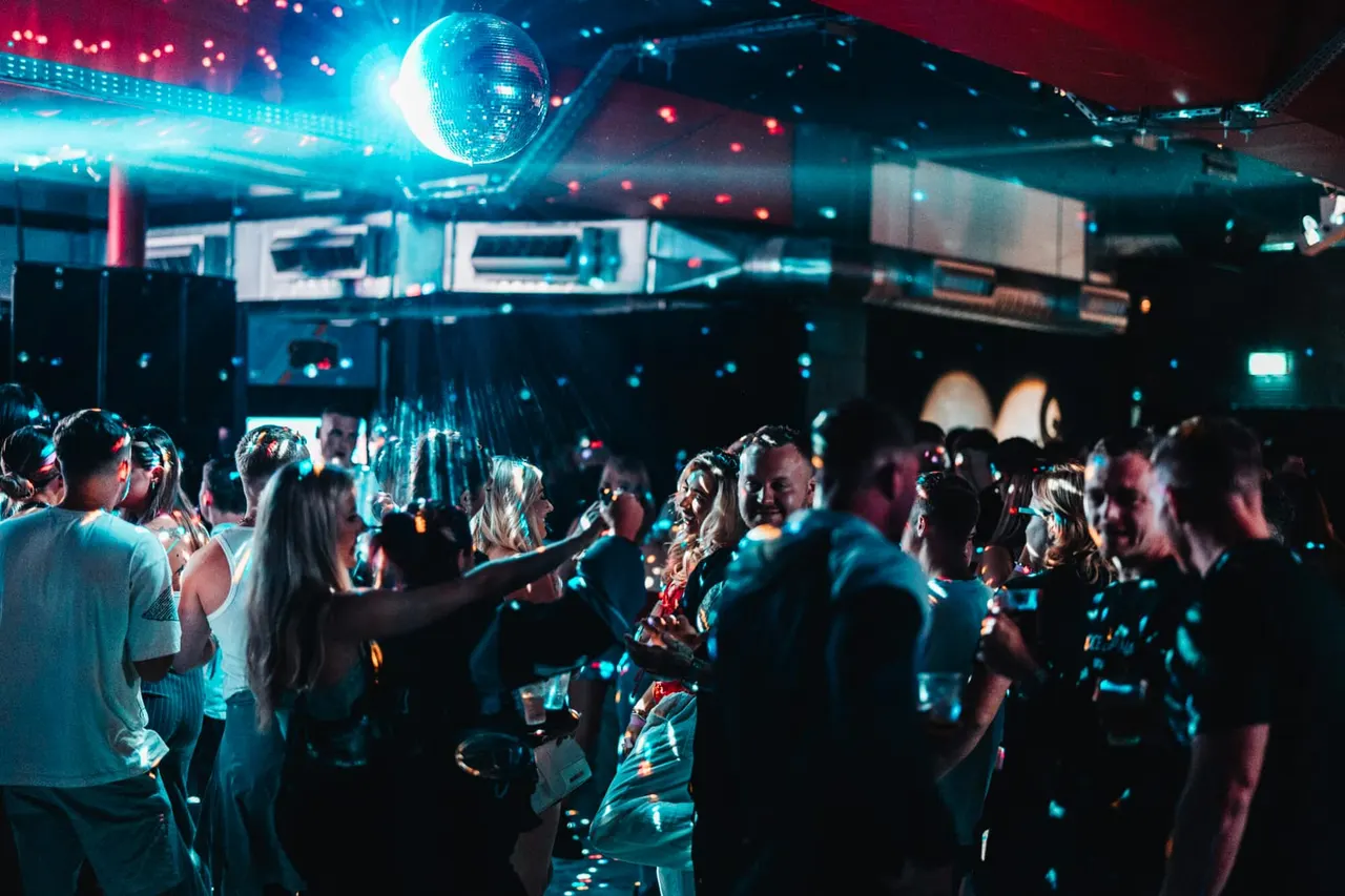 Crowded nightclub dancefloor with disco ball overhead and neon pink and blue lighting, guests dancing and socialising.