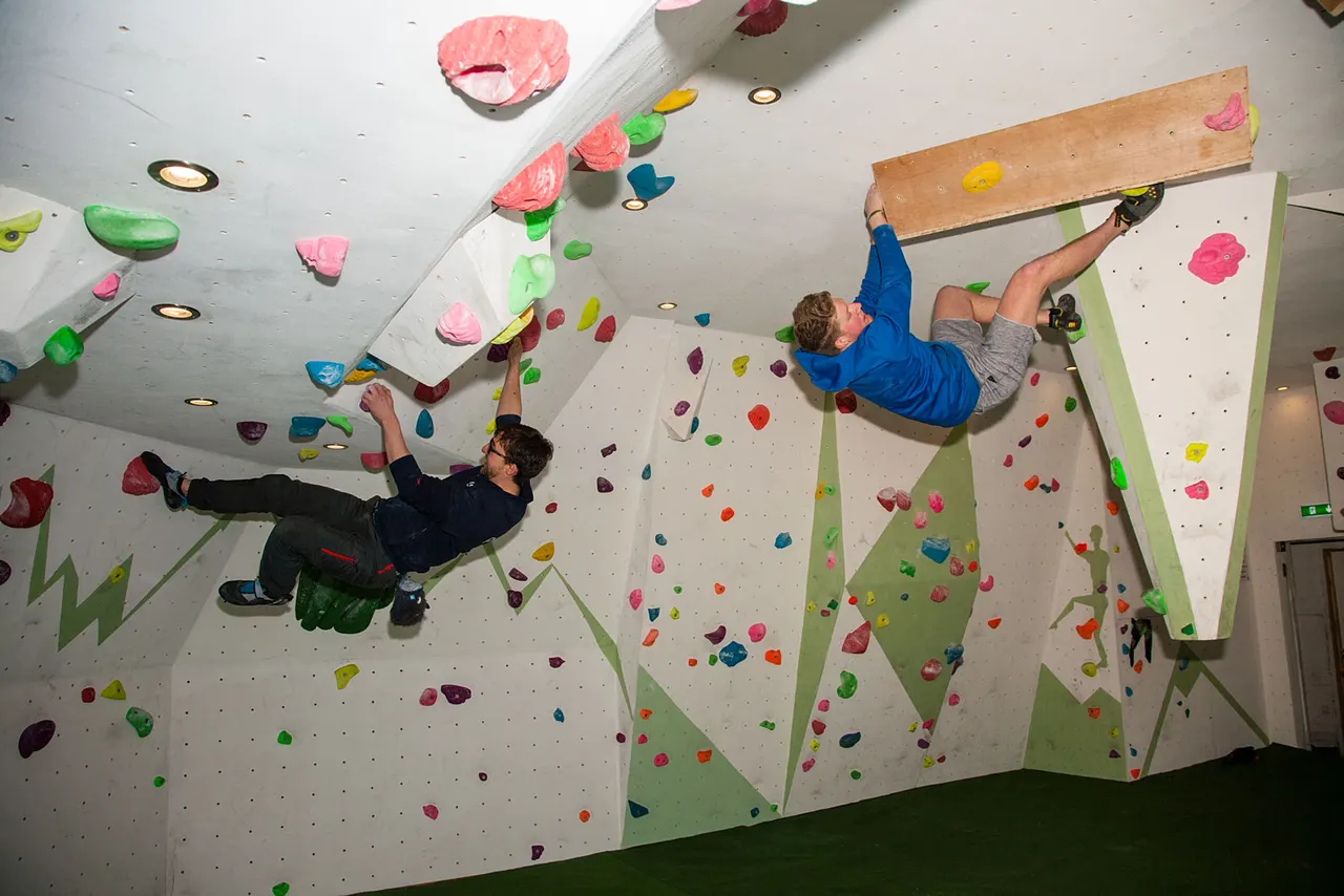 Two climbers ascending indoor climbing walls with coloured holds, one on an overhang and one on a vertical section.